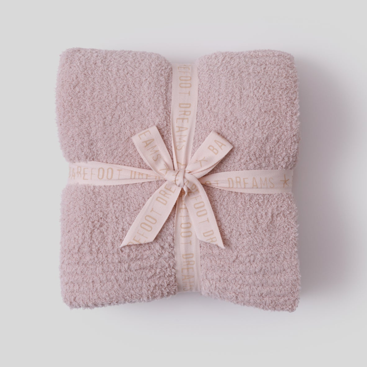 A folded cream-colored Barefoot Dreams CozyChic throw blanket tied with a branded ribbon, presented as a luxury relaxation gift.